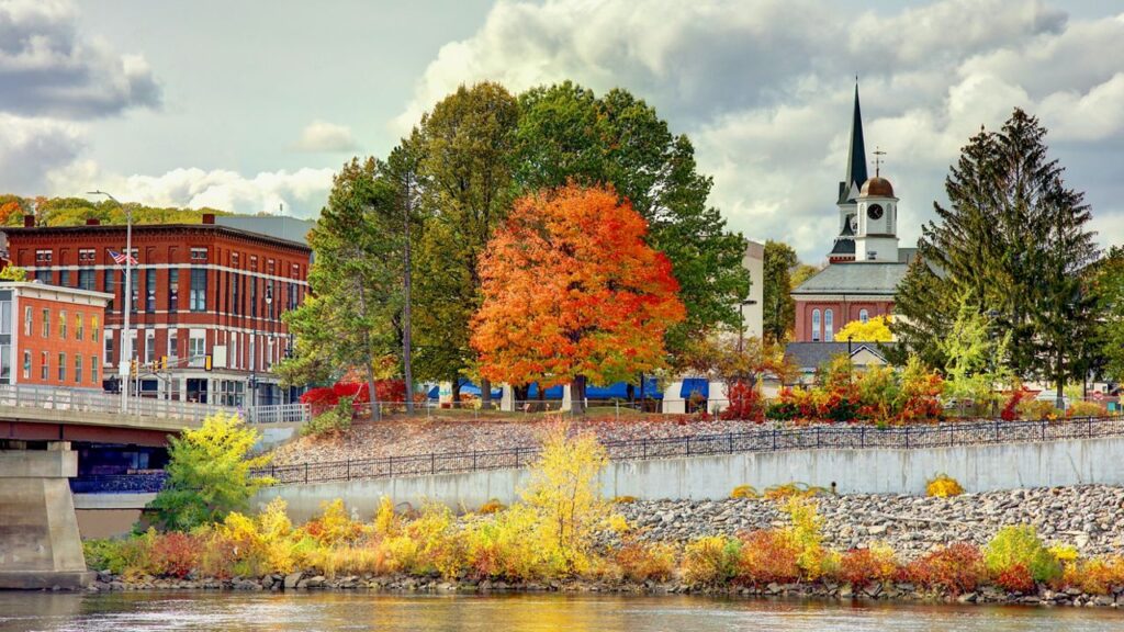 Vibrant autumn scene in Auburn, Maine featuring a colorful tree-lined riverbank, historic brick buildings, and a church steeple rising in the background—capturing the city’s charm and seasonal beauty.