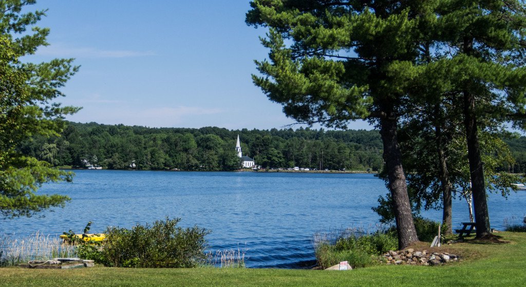 Peaceful view of China Lake in Maine with a white steeple peeking through trees in the distance, symbolizing the town’s quiet beauty and deep-rooted community values.