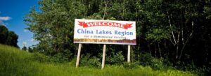 Welcome to China, Maine" roadside sign surrounded by trees on a clear summer day, representing the rural charm and welcoming spirit of the town.