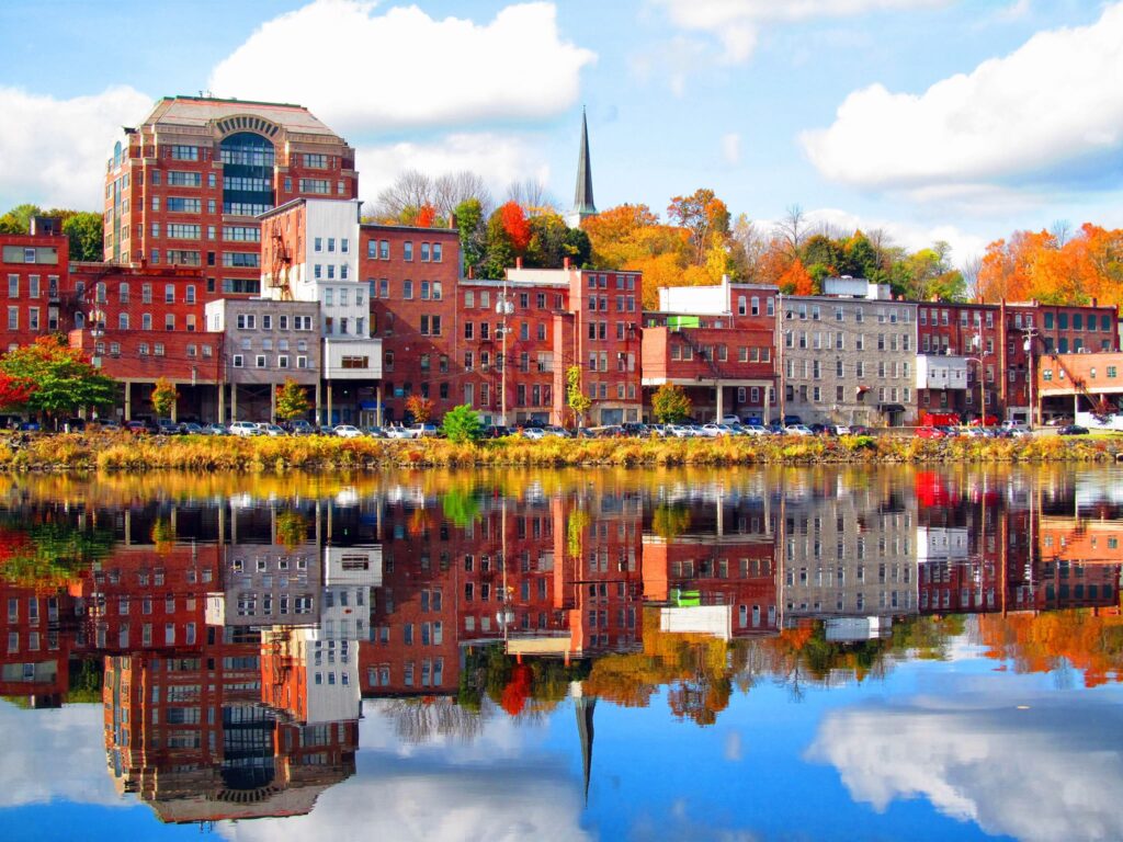 Colorful historic buildings along the Kennebec River in downtown Augusta, Maine, reflected in calm water with vibrant fall foliage and a church steeple in the background.