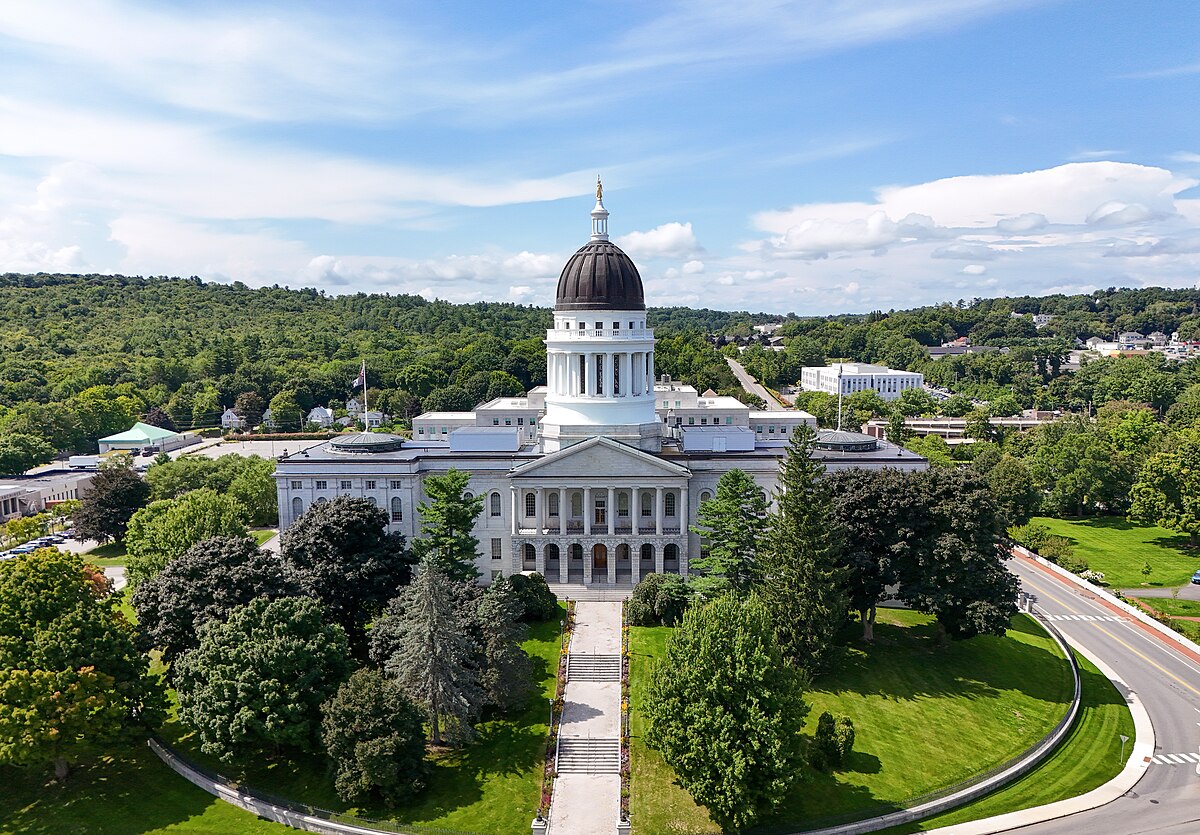 Aerial view of the Maine State House in Augusta, surrounded by green trees and hills under a partly cloudy sky.