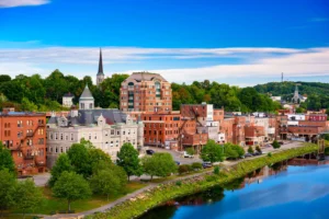 Downtown Augusta, Maine overlooking the Kennebec River, with historic brick buildings, green trees, and the Maine State House dome in the distance under a bright blue sky.
