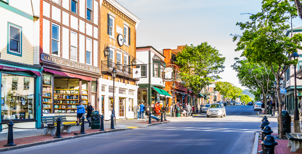 Vibrant downtown street in Bar Harbor, Maine, lined with colorful storefronts, a clock tower, and people enjoying shops and cafes on a sunny day.