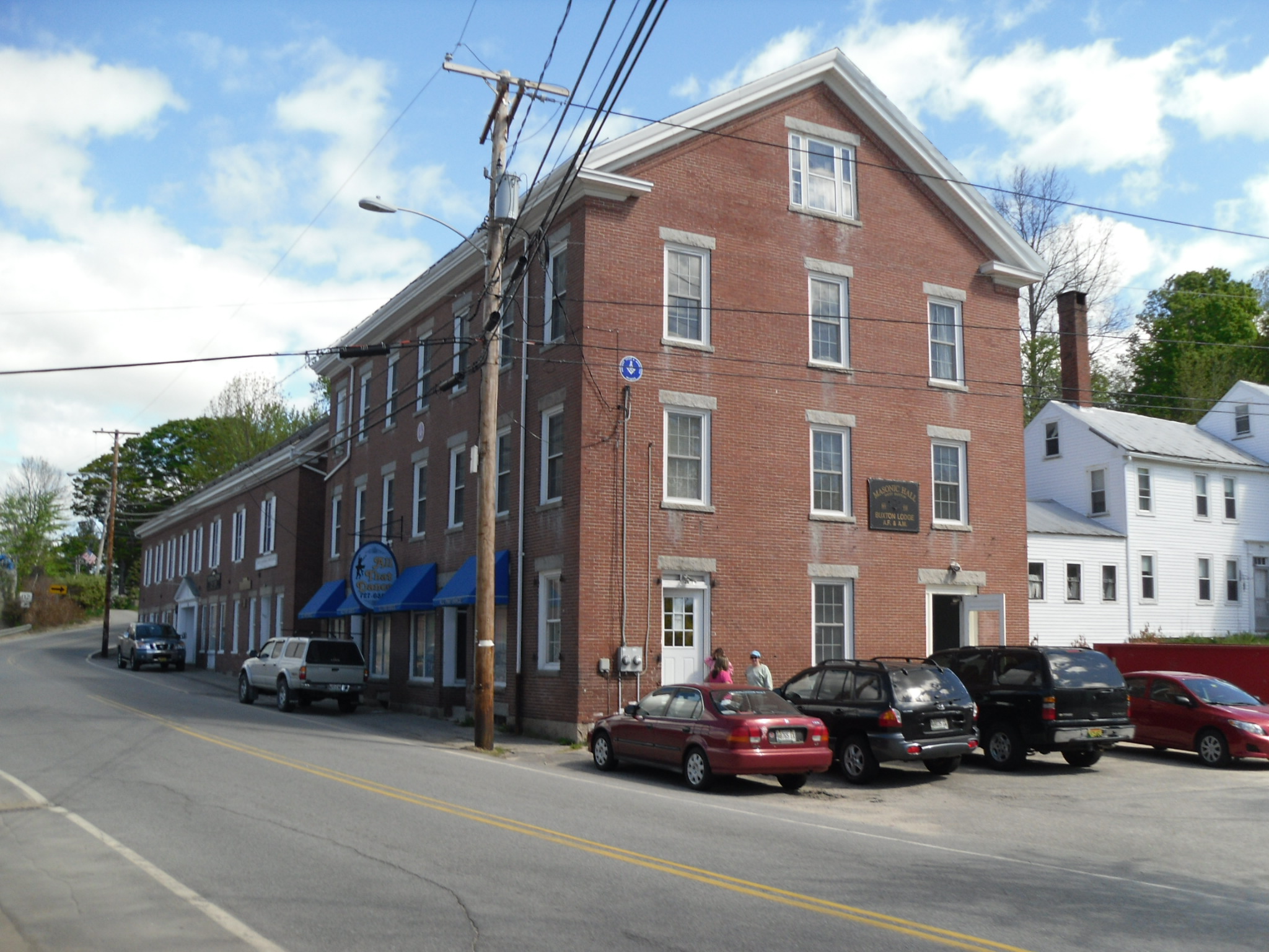 Historic brick building and local businesses along Main Street in Buxton, Maine with cars parked out front under a bright sky.