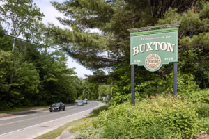 Welcome sign at the roadside entrance to Buxton, Maine surrounded by trees and greenery.