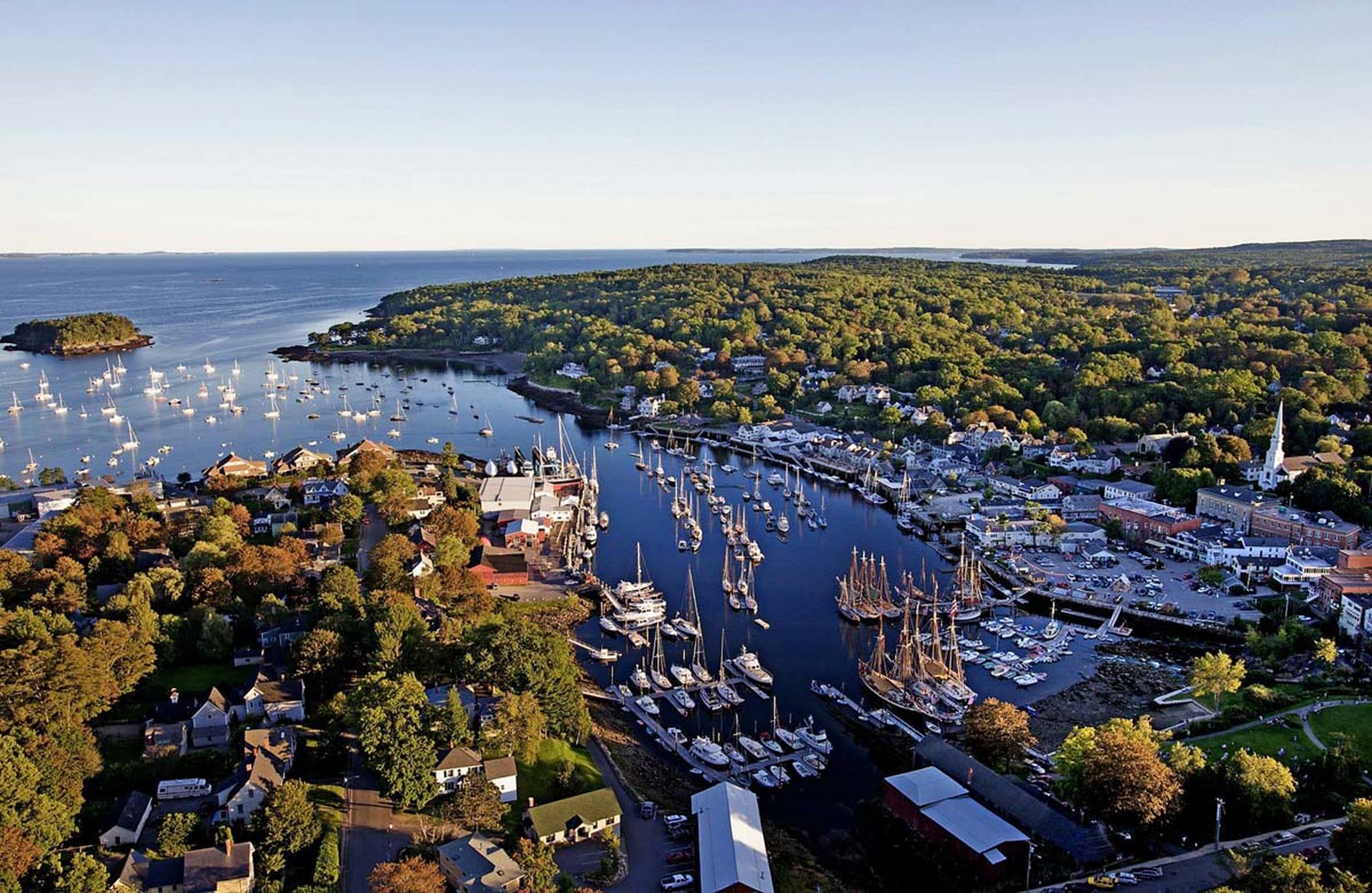 Aerial view of Camden Harbor, Maine at sunset, with sailboats docked in the marina, downtown buildings nearby, and lush green hills in the background.