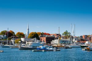 View of Camden Harbor on a clear day, with sailboats docked in the foreground and the town’s buildings and hills visible in the distance.