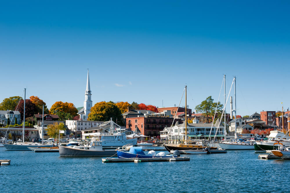 View of Camden Harbor on a clear day, with sailboats docked in the foreground and the town’s buildings and hills visible in the distance.