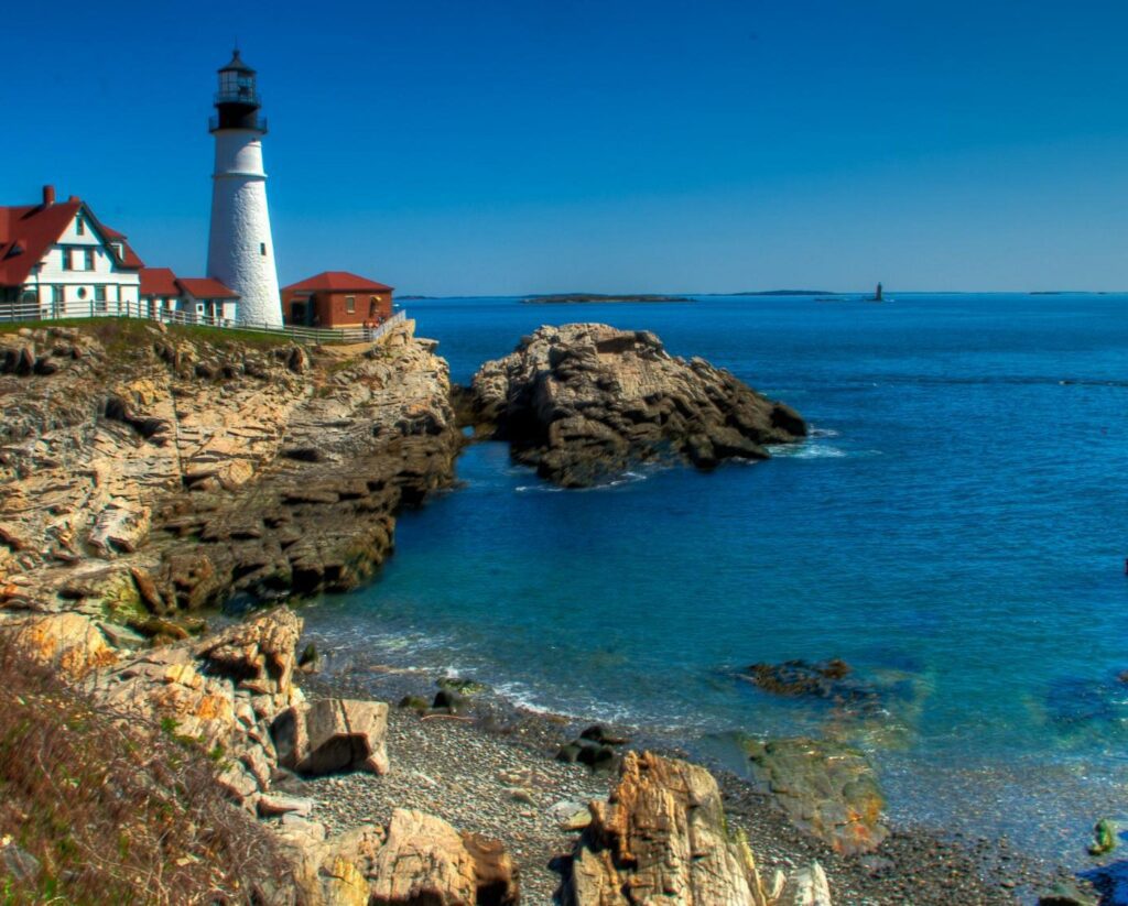 Rocky shoreline view of Cape Elizabeth, Maine, with a lighthouse perched on cliffs beside bright blue ocean waters under a clear sky.