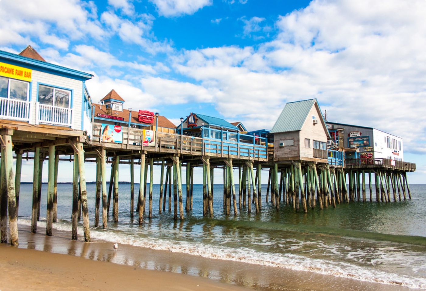 View of Cape Elizabeth pier with colorful seaside buildings and reflections on the water, under a partly cloudy sky.