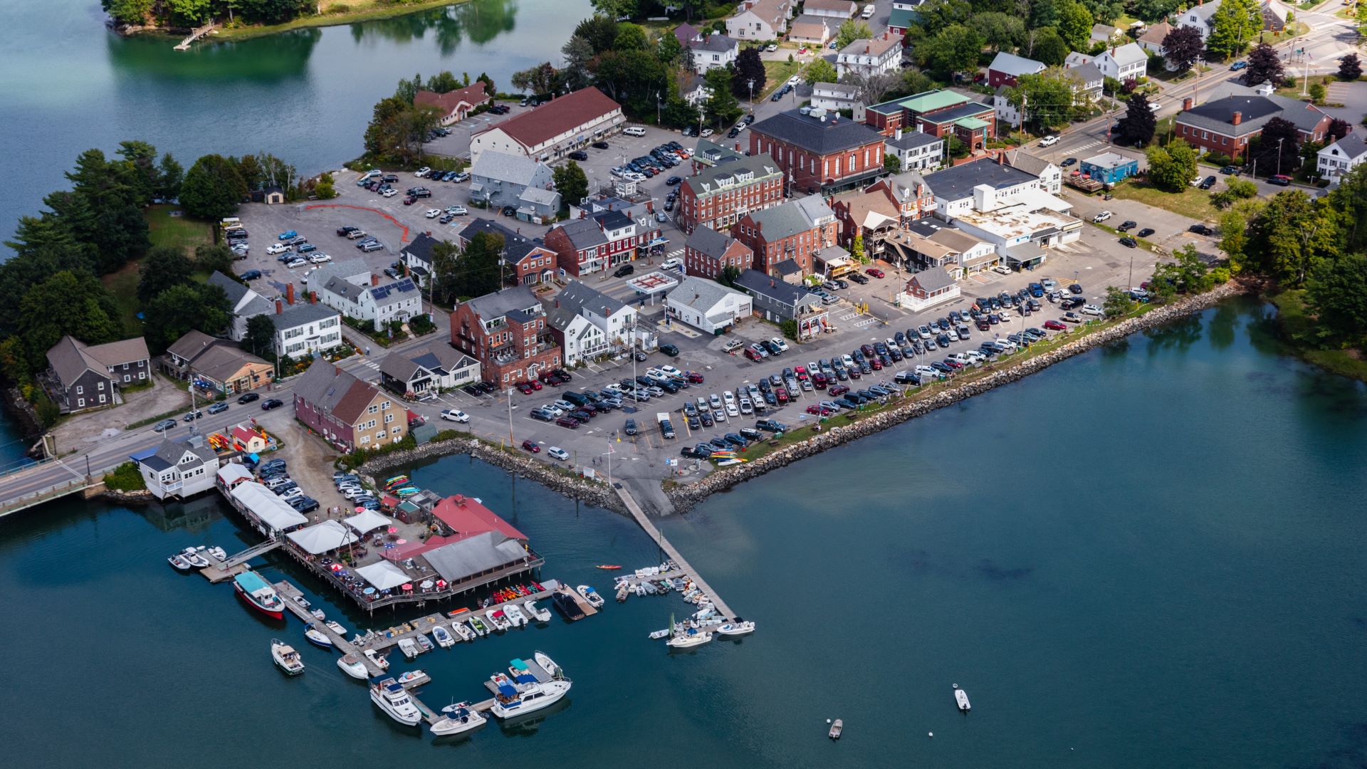 Aerial view of Damariscotta, Maine, showing a waterfront marina with boats, red-roofed buildings, parking lots, and historic downtown architecture surrounded by trees and calm waters.