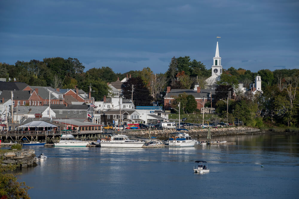 Boats docked along the calm Damariscotta River with historic waterfront buildings and a white steeple rising in the background under a partly cloudy sky.