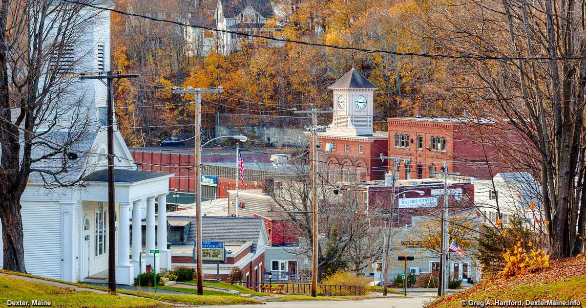 Downtown Dexter, Maine in autumn, showing historic buildings, a clock tower, utility poles, and colorful fall trees.