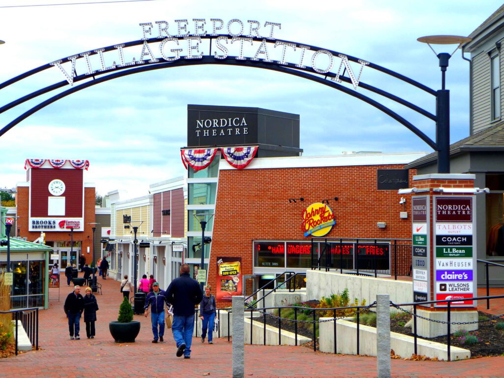 Pedestrian shopping plaza beneath the Freeport, Maine welcome arch, with local shoppers walking between brick storefronts and outlet shops.