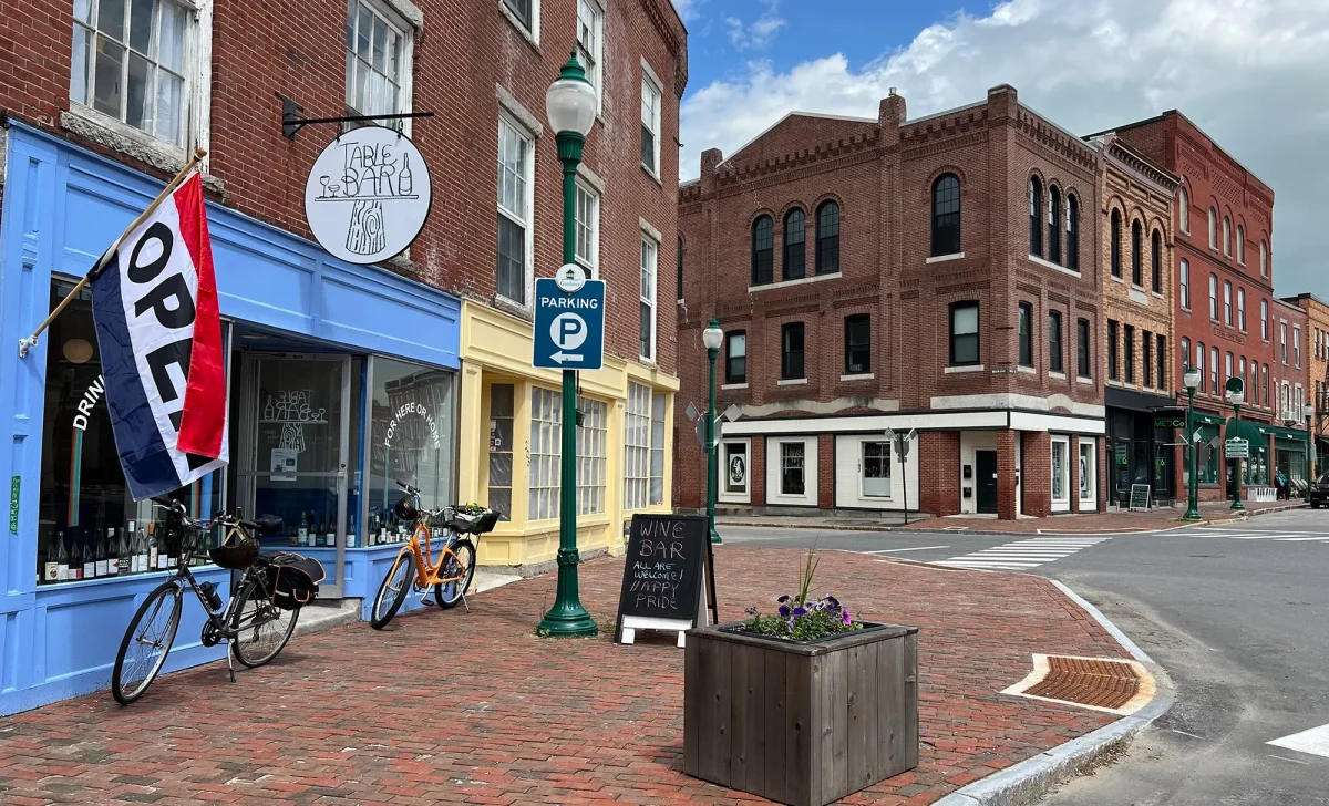 Downtown Gardiner, Maine storefronts with an open flag, bicycles, and historic brick buildings on a sunny day.