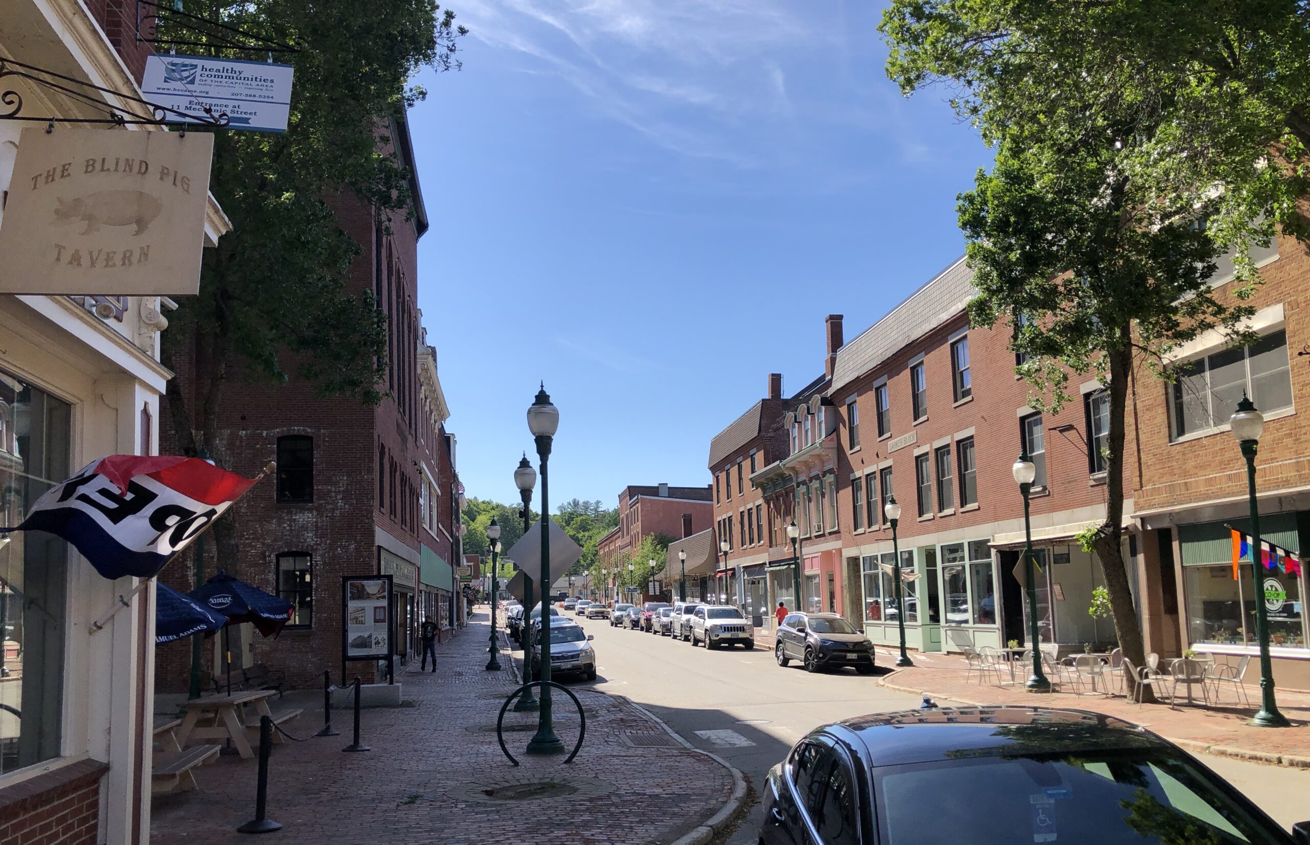Downtown Gardiner, Maine on a sunny day, featuring historic brick buildings, tree-lined sidewalks, and local shops along Water Street.